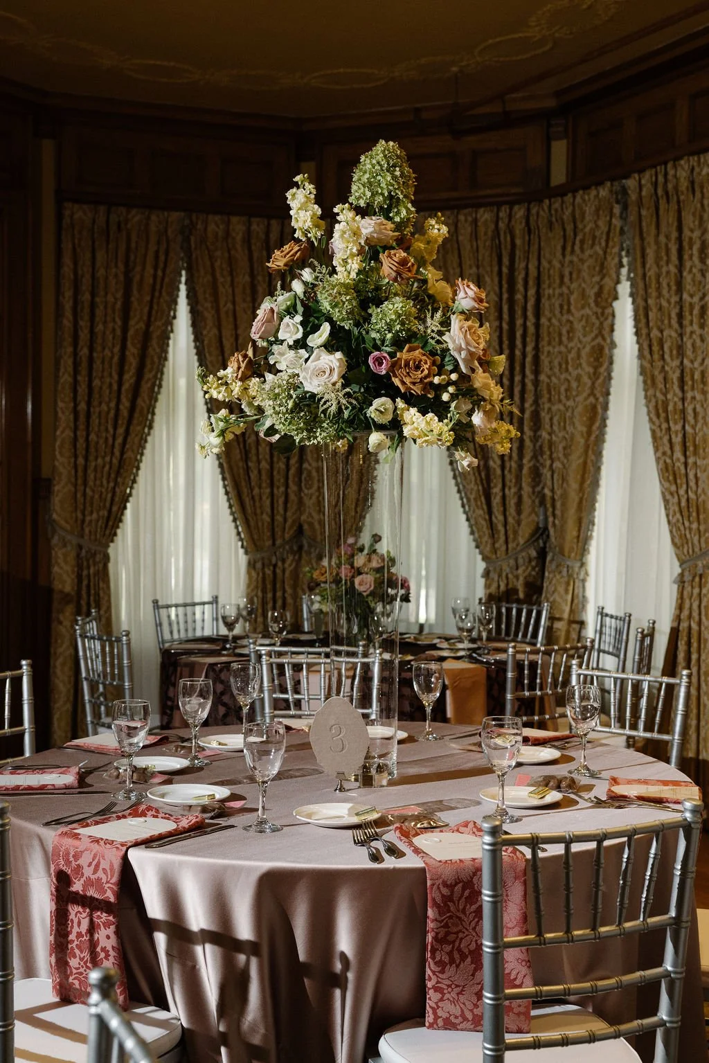 Elegant banquet table with a tall floral centerpiece in a formal dining room with gold curtains.