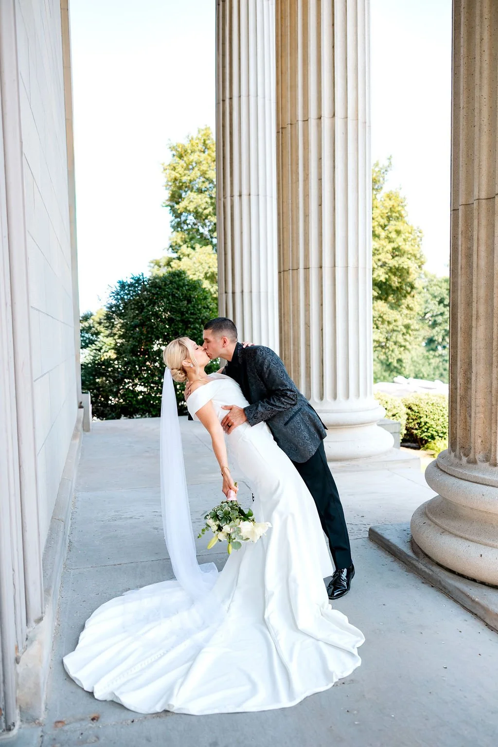A newlywed couple sharing a kiss outside on the steps of a building with large columns, greenery in the background, and bright sunlight.