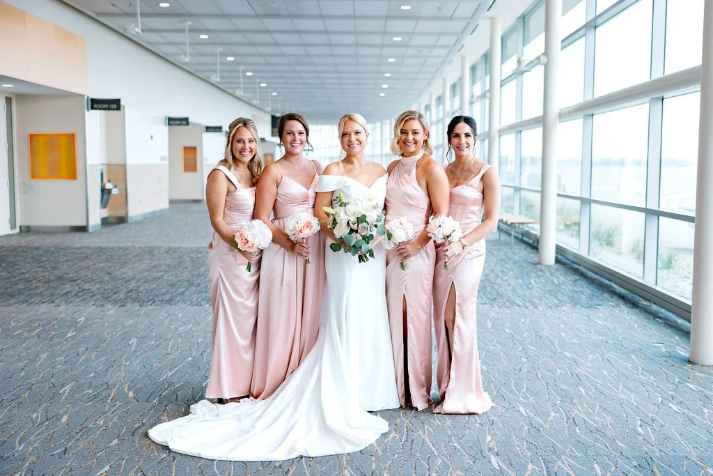 Group of five women in an indoor airport terminal, dressed in wedding attire, with the bride in a white gown and four bridesmaids in matching blush pink dresses, holding bouquets of white flowers.