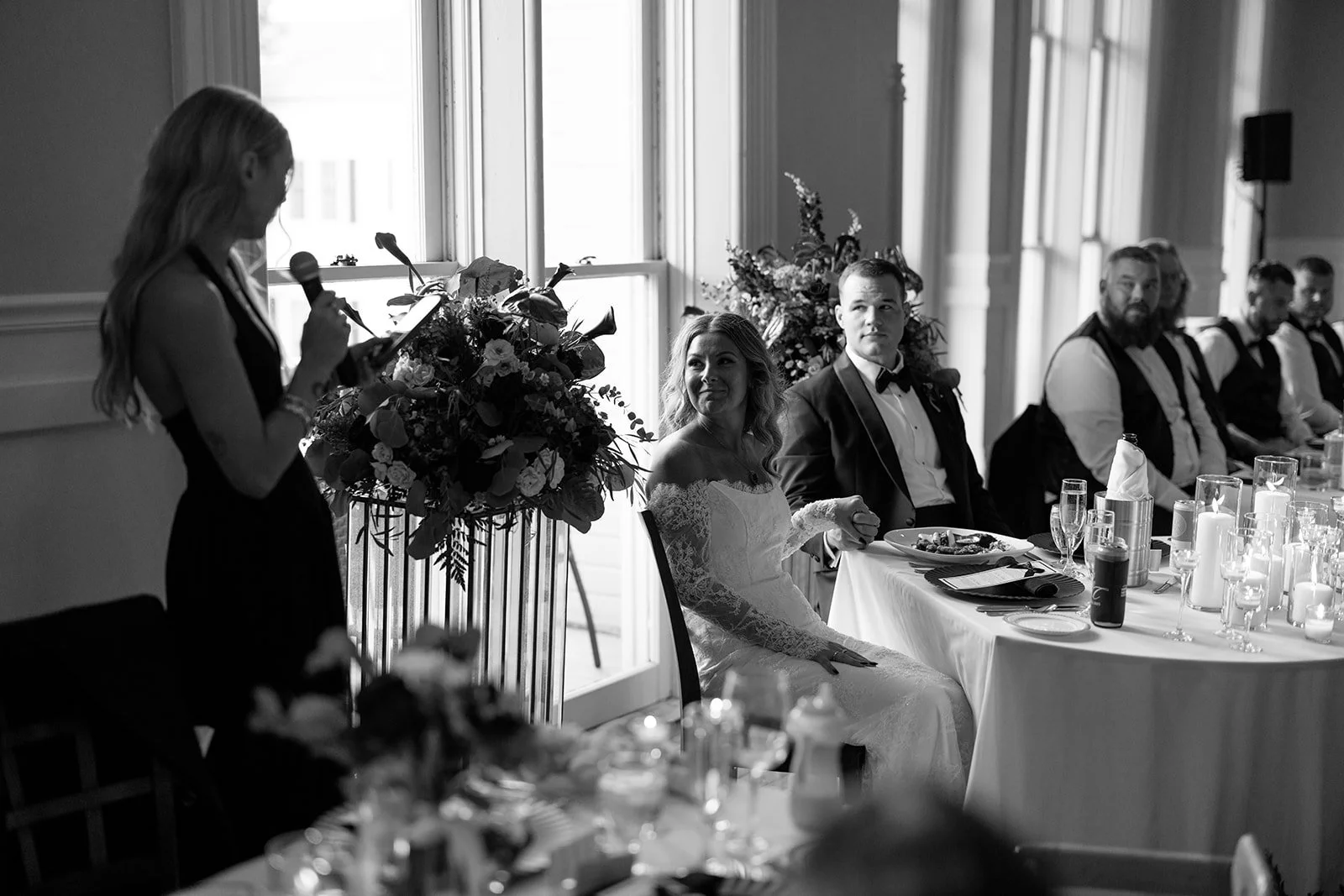 A wedding reception scene with a bride and groom sitting at a table, listening to a woman giving a speech. The setting is elegant with floral arrangements and candles on the table.