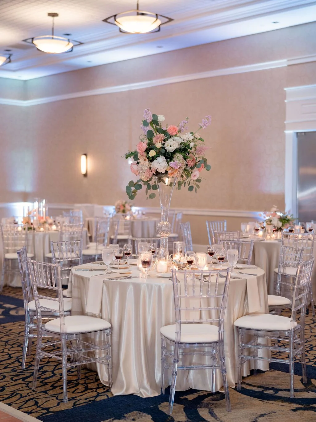 Wedding reception setup with round tables, clear chairs, elegant floral centerpieces, and soft lighting.