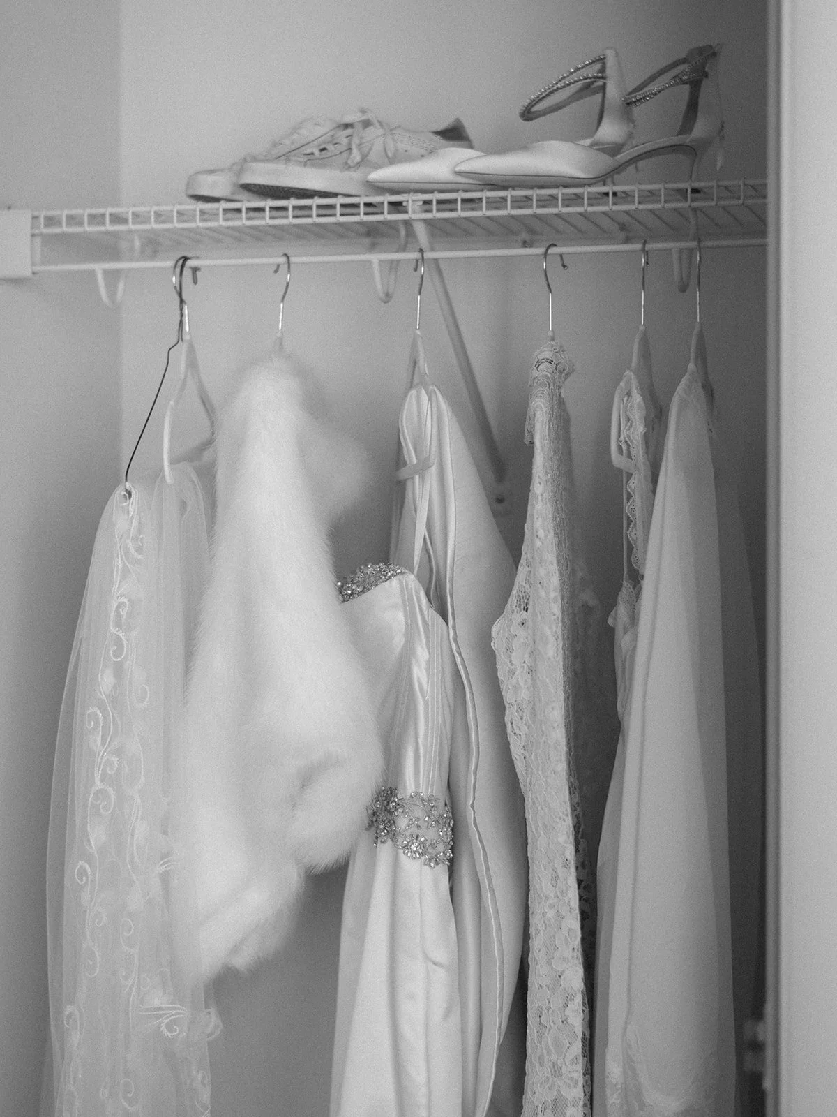 A closet shelf with a pair of high-heeled shoes with ankle straps, a white fur stole, and several hanging wedding dresses with lace and embellishments.