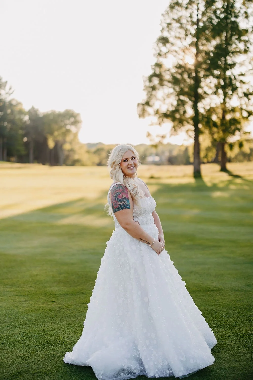 A woman with blonde hair and tattoos is wearing a white wedding dress and is standing outdoors on a grassy field during sunset, smiling at the camera with trees in the background.