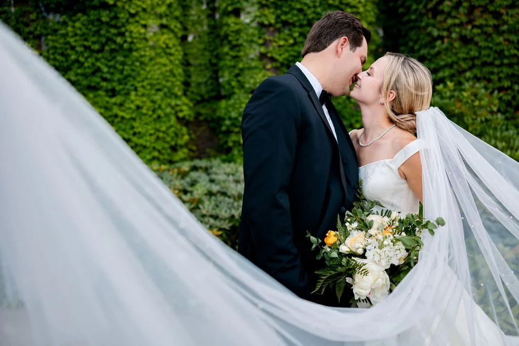 A newlywed couple sharing a kiss outdoors, with the bride holding a bouquet of white and yellow flowers, and the couple dressed in wedding attire.