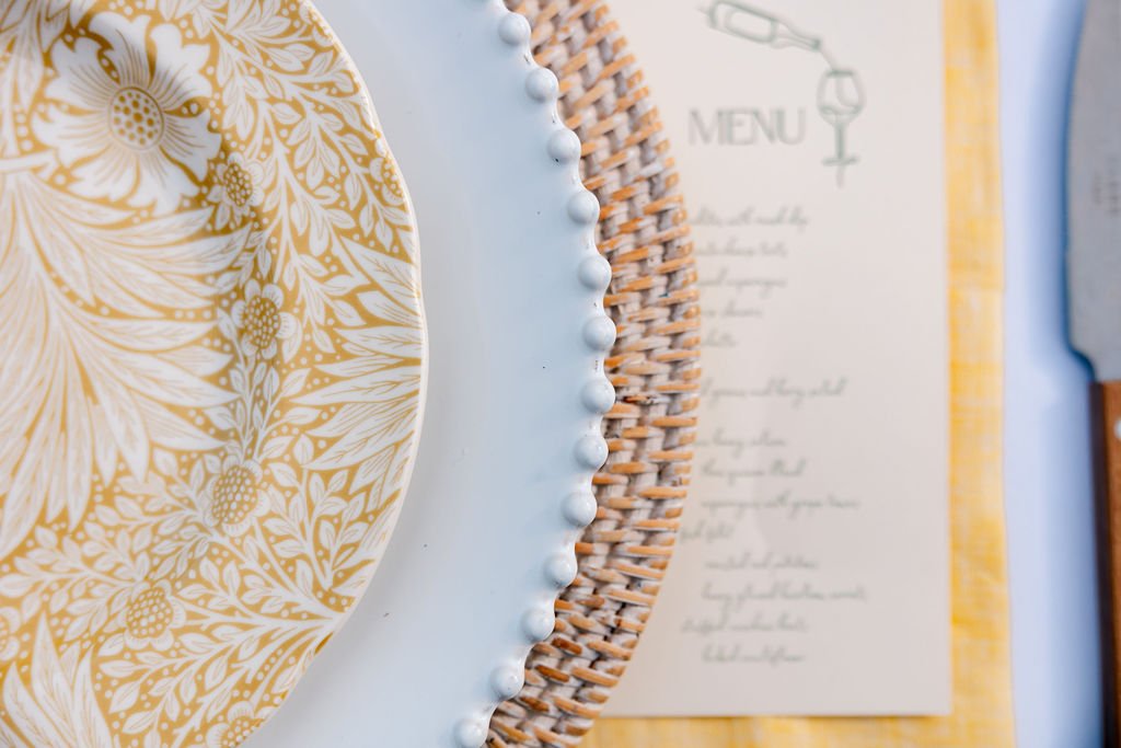 Close-up of a decorative white plate with a yellow floral pattern on a dining table, with a menu and a woven placemat in the background.