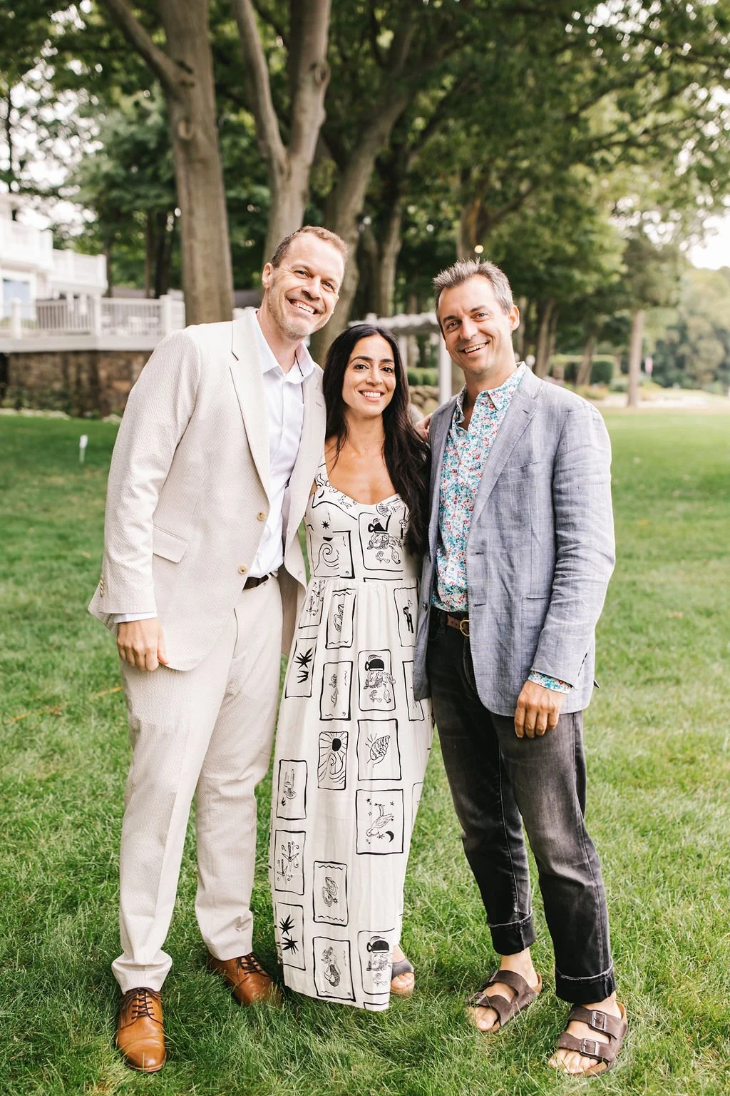 Three people standing together outdoors on a lush green lawn with trees in the background, smiling at the camera.