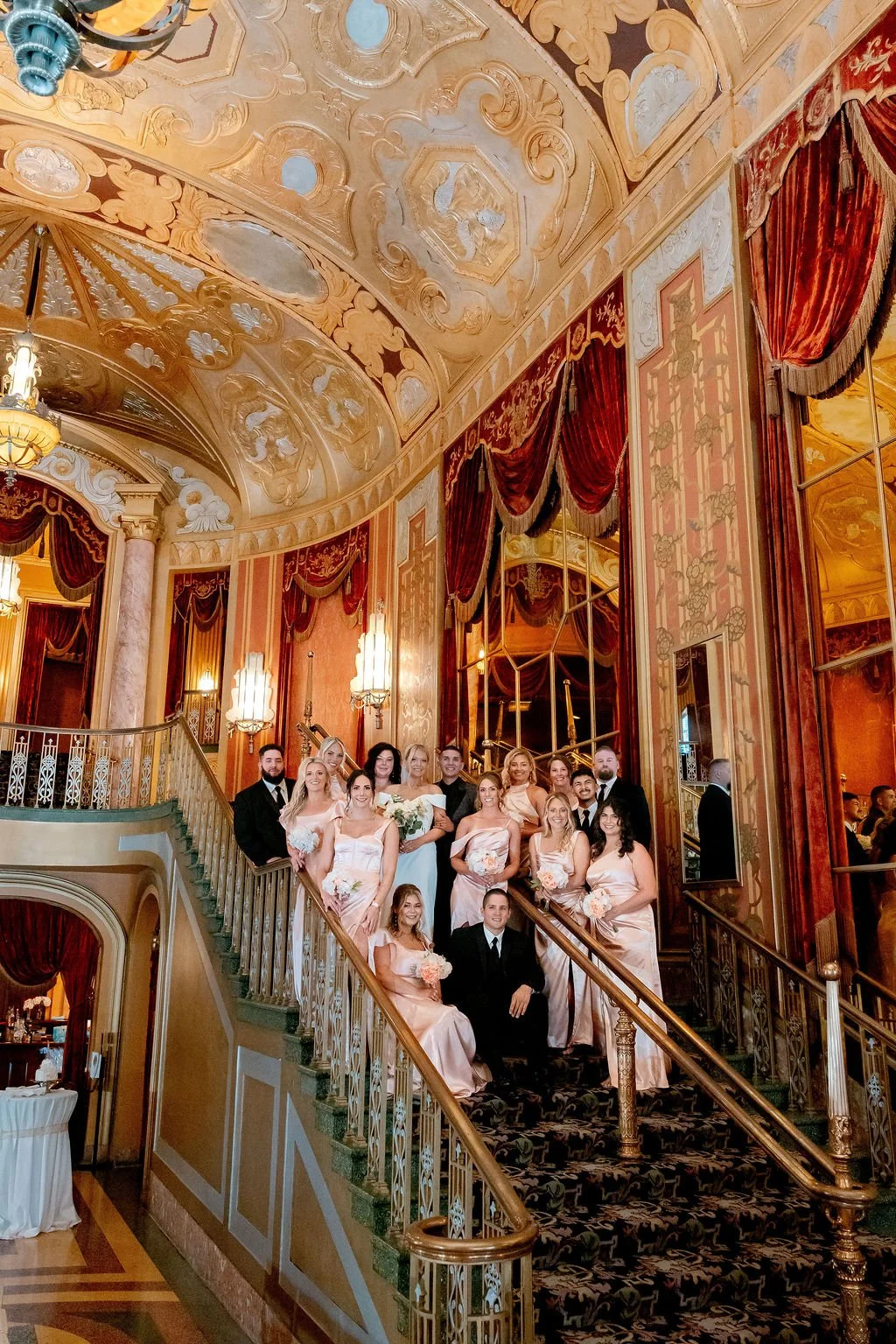 A group of people dressed in formal attire, including bridesmaids in peach-colored dresses and groomsmen in black tuxedos, standing on a grand staircase inside an ornate, luxurious hall with gold and red decorations, stained glass ceiling, and elabor