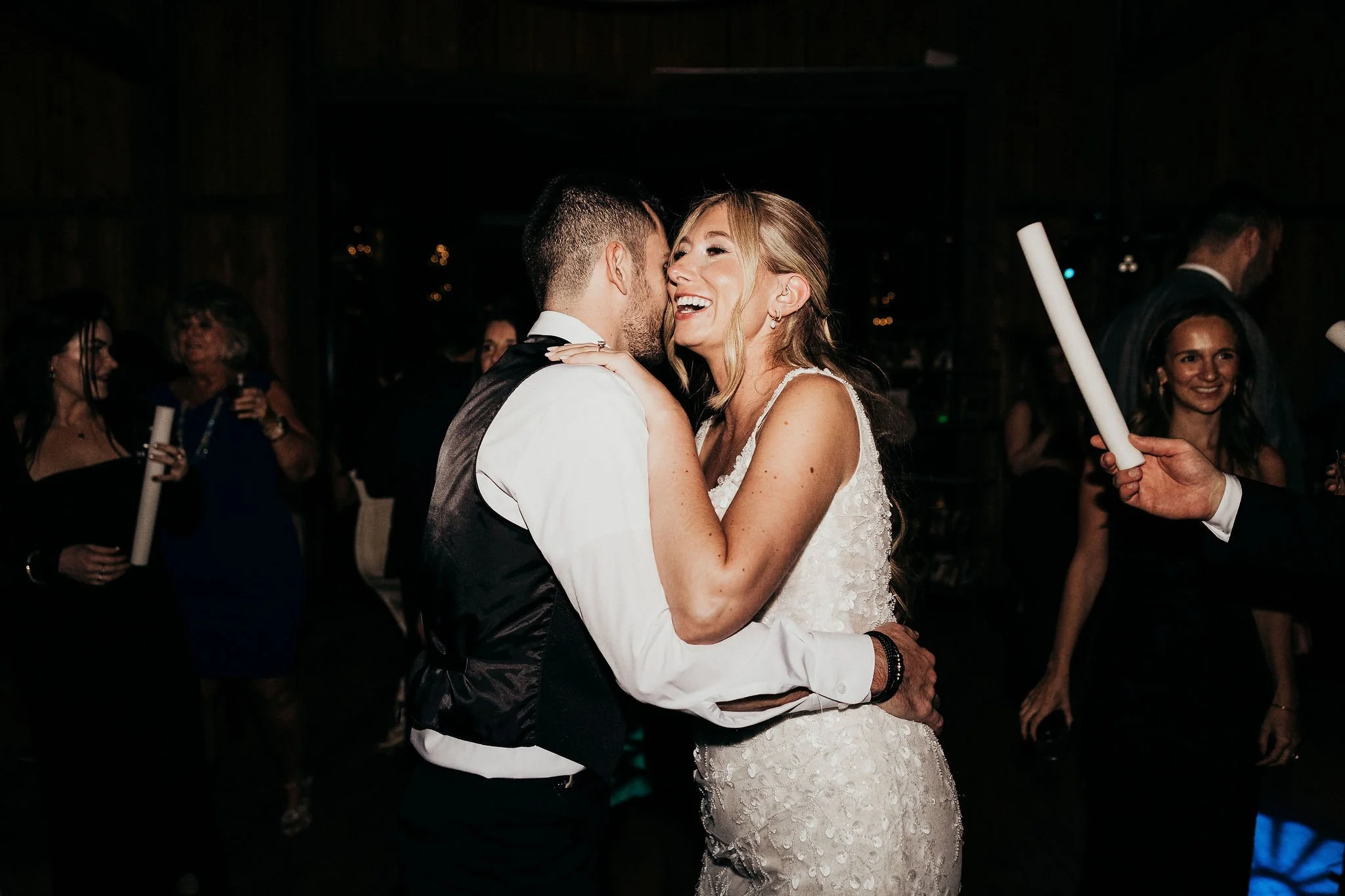 A newlywed couple sharing a dance at their wedding reception, surrounded by guests in a dimly lit venue.