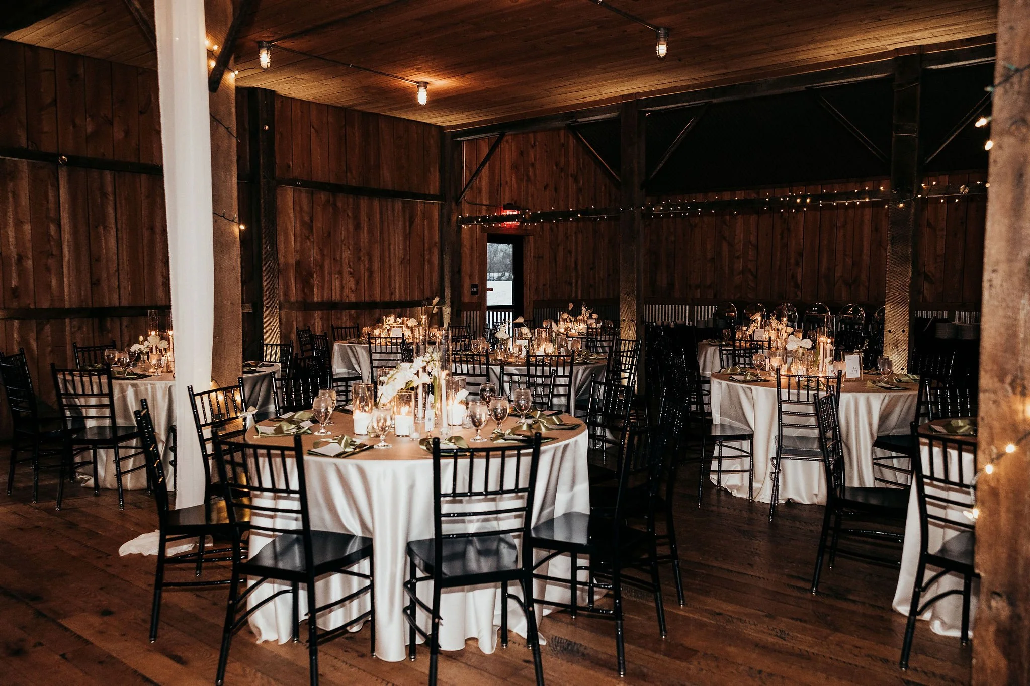 Elegant indoor event space with round tables covered in white tablecloths, decorated with flowers, candles, and glassware. The ceiling and walls are made of wood, and warm string lights hang across the space, creating a cozy ambiance.
