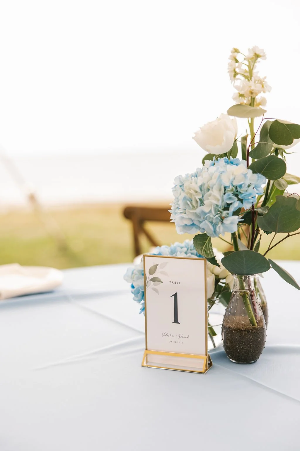 A wedding table centerpiece featuring a glass vase with blue and white hydrangeas, white roses, and green leaves, accompanied by a white table number card with gold accents reading 'Table 1' and the date '8.23.2025'.