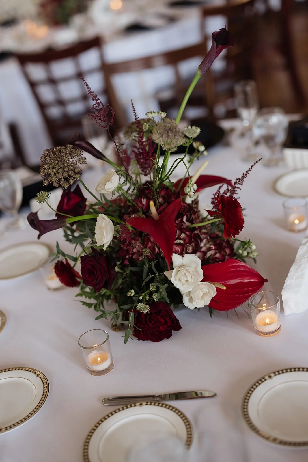 Elegant table centerpiece with red, white, and dark purple flowers, surrounded by small candles in glass holders on a white tablecloth with fine china and silverware.
