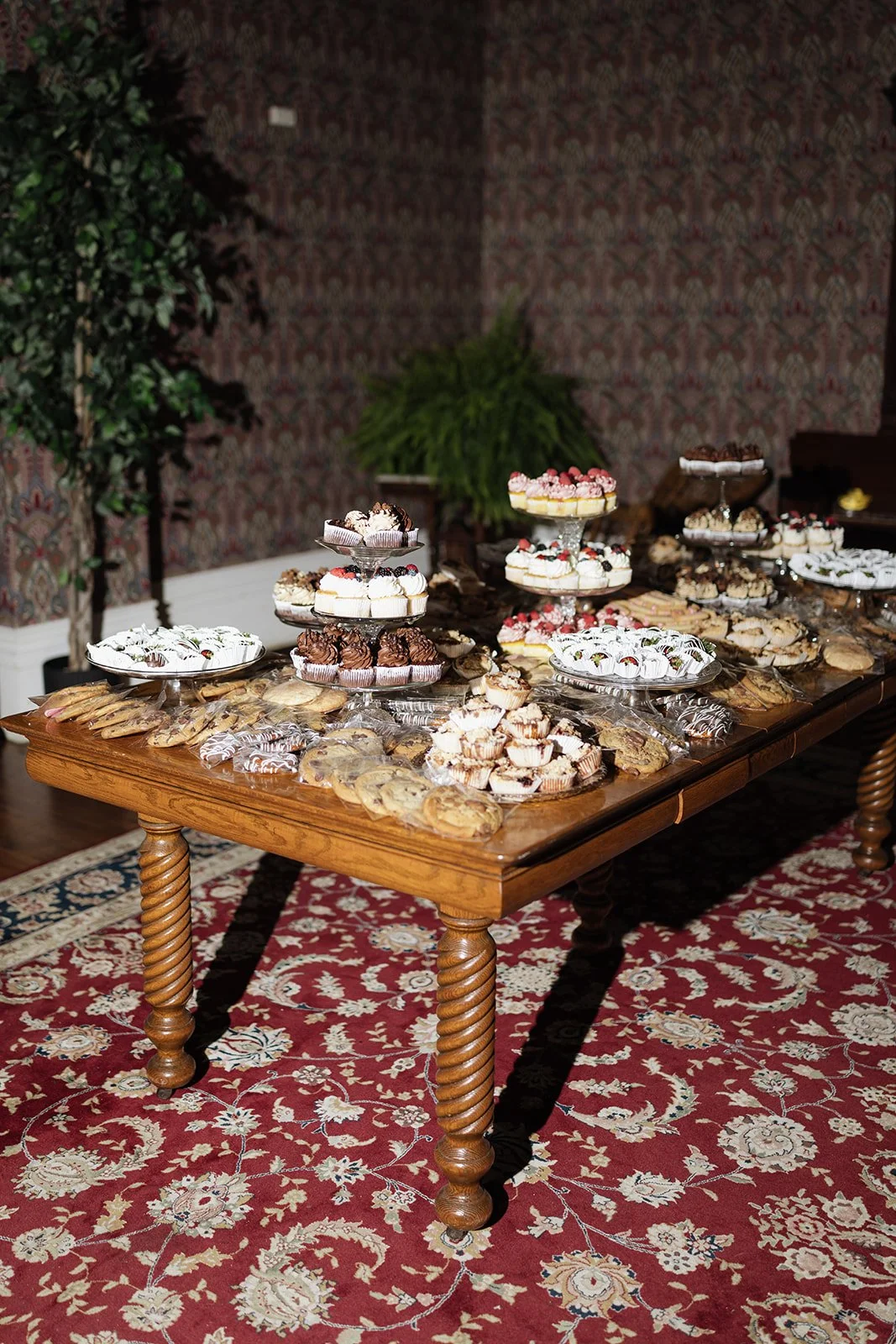 A wooden table filled with assorted cookies and cupcakes decorated with frosting and berries, set on a red patterned rug in a dimly lit room with patterned wallpaper and green plants in the background.