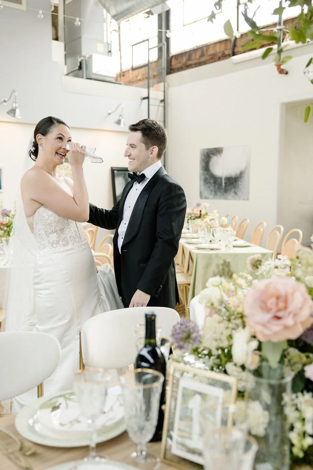 Happy bride with a tissue, smiling as groom in tuxedo gently touches her face during wedding reception with decorated tables and floral centerpieces in the background.