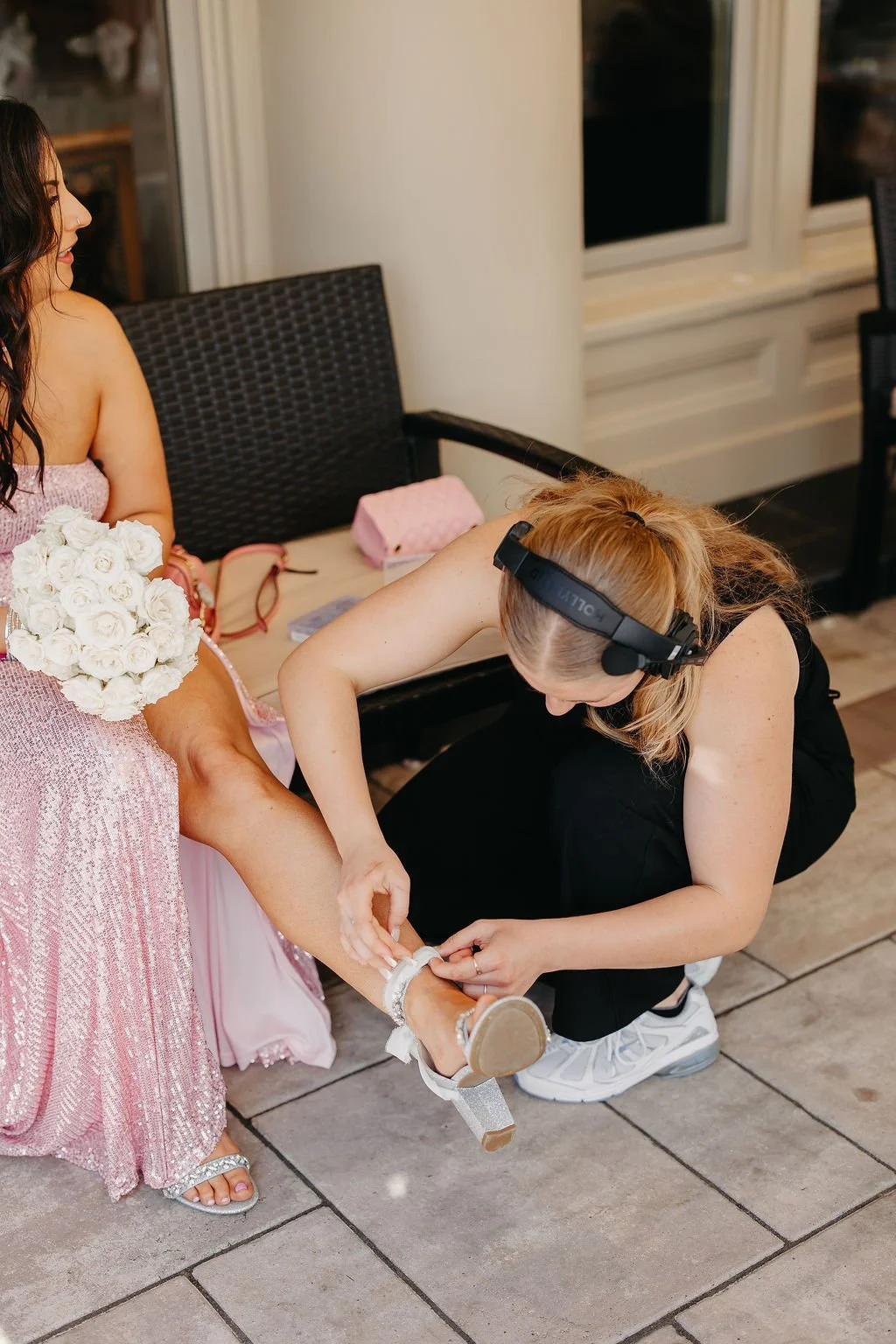 A woman dressed in a pink, sparkly gown is sitting on a bench, holding a bouquet of white roses. A woman wearing black pants, white sneakers, and a headset is kneeling and helping her put on a silver, glittery high heel shoe.