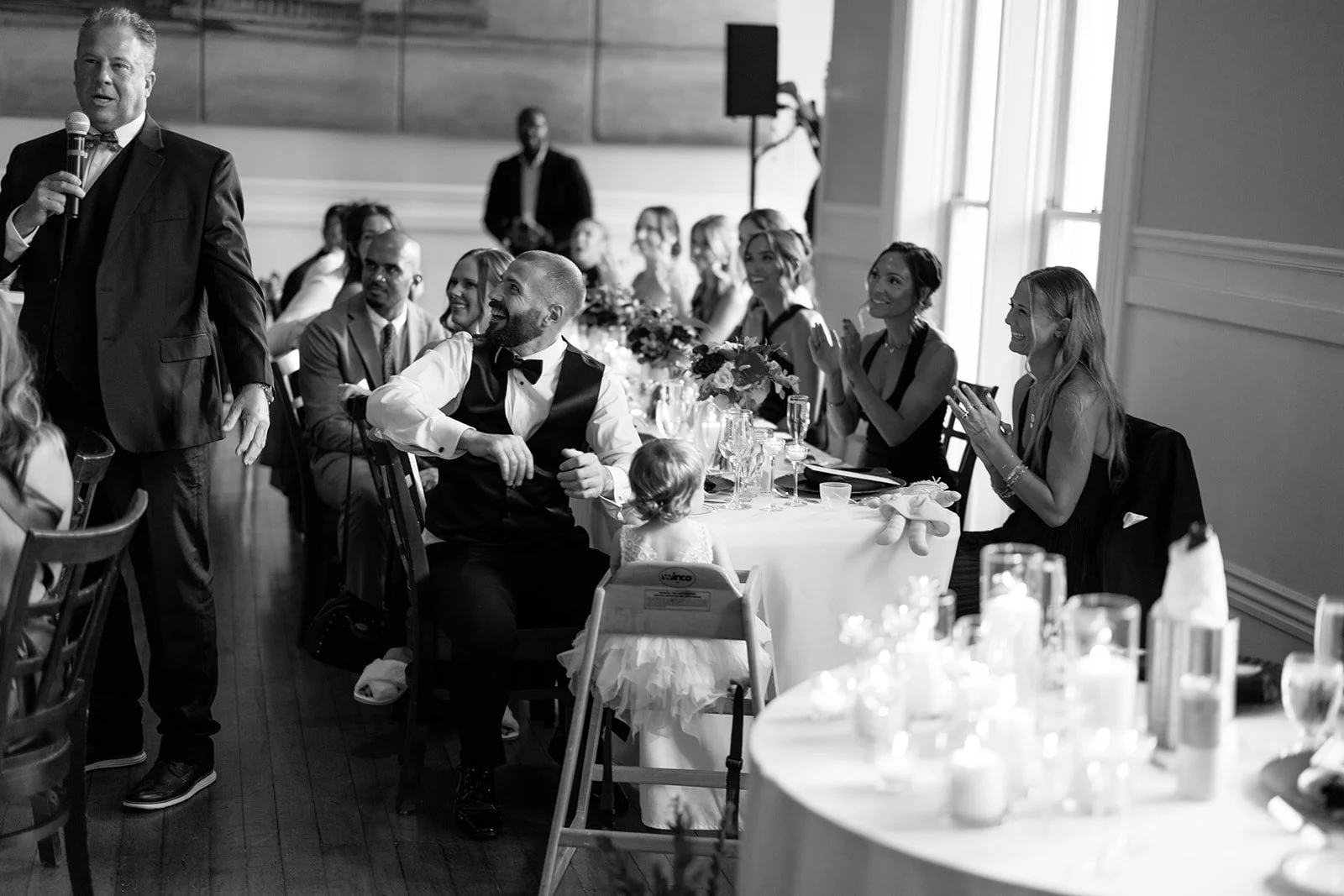 A man in a tuxedo giving a speech at a formal gathering, with guests seated at a round table, smiling and clapping, in a well-lit room with large windows.