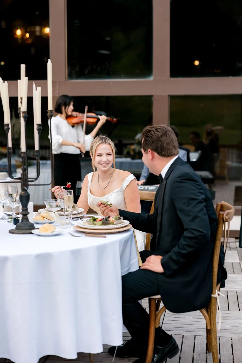 A romantic dinner at a wedding reception with a bride and groom, a woman with blonde hair and a man with brown hair, sitting at a table with elegant place settings, wine glasses, bread rolls, and a candelabra. A violinist plays in the background, and