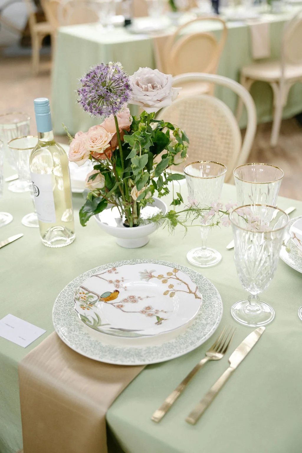 Elegant dining table set with floral centerpiece, porcelain plates featuring bird and cherry blossom design, gold-rimmed glassware, gold cutlery, and a pale green tablecloth at a formal event or wedding reception.