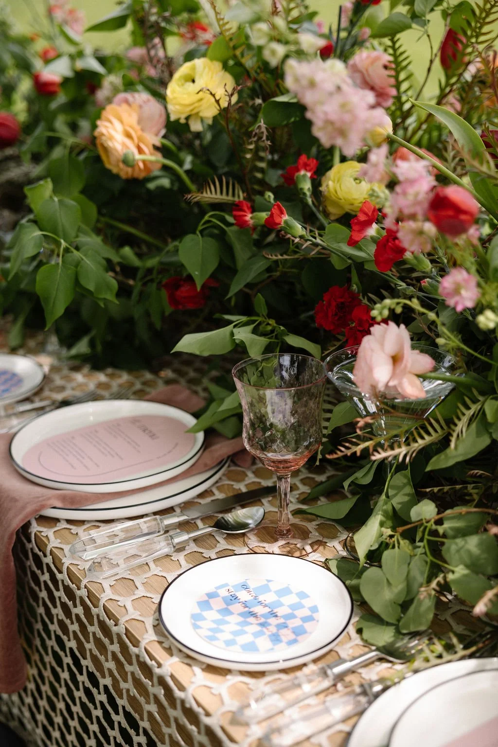 A decorated table with plates, glasses, utensils, and pink menus, surrounded by colorful flowers and greenery for a celebration or event.