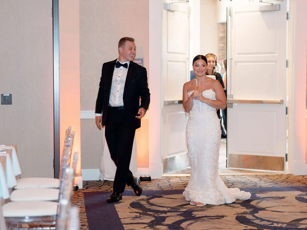 Bride in wedding dress holding a drink, smiling as she enters a reception, groom in tuxedo walking nearby, reception hall with decorated tables.