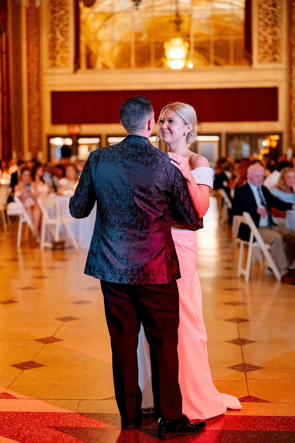 A couple dressed in formal attire dancing at a wedding reception, with guests seated at tables in the background.