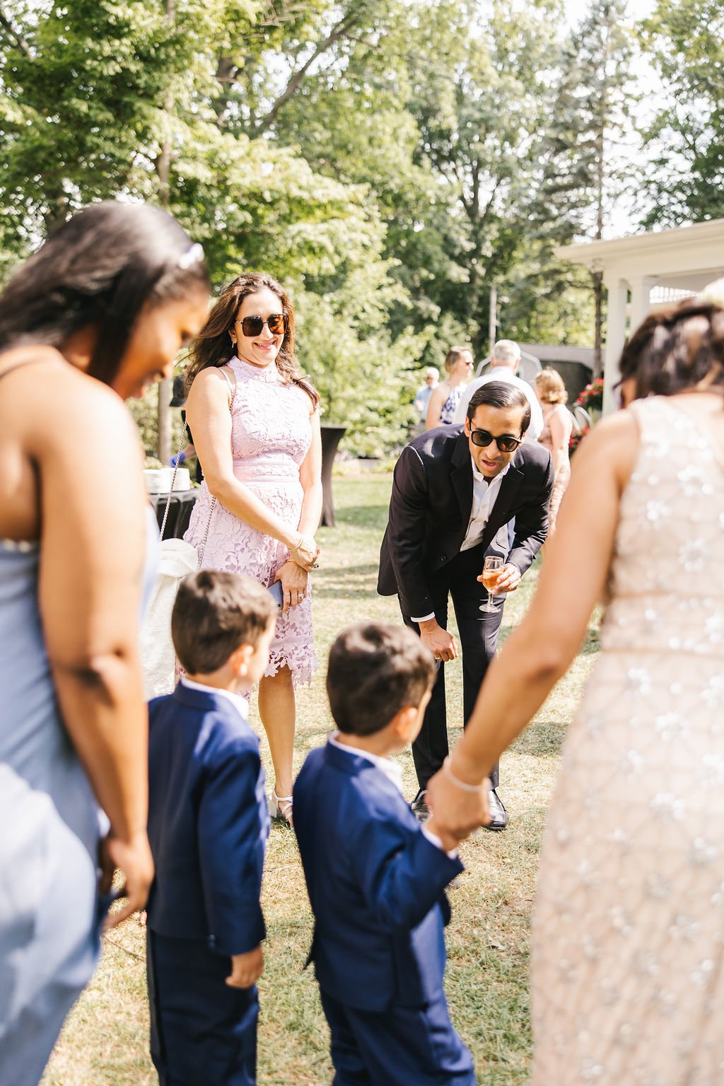 Group of people at an outdoor event, children in suits, adults talking, one man bending down with a drink, woman in a pink dress, woman in a beaded dress, and a woman in a gray dress, on a sunny day in a garden setting.