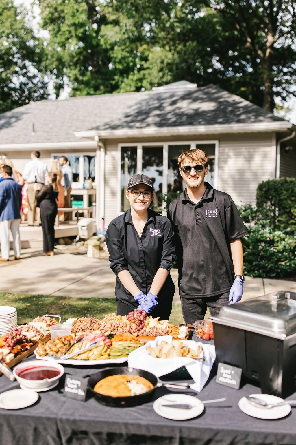 Two smiling catering staff members, a woman and a man, standing behind a table with a variety of food at an outdoor event, with a house and guests in the background.