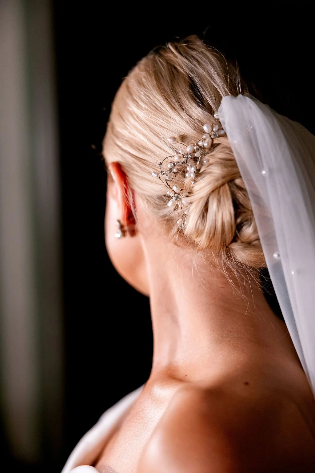 A bride's updo hairstyle adorned with a pearl and wire hair accessory, with a white veil attached, and her ear with a pearl earring visible.