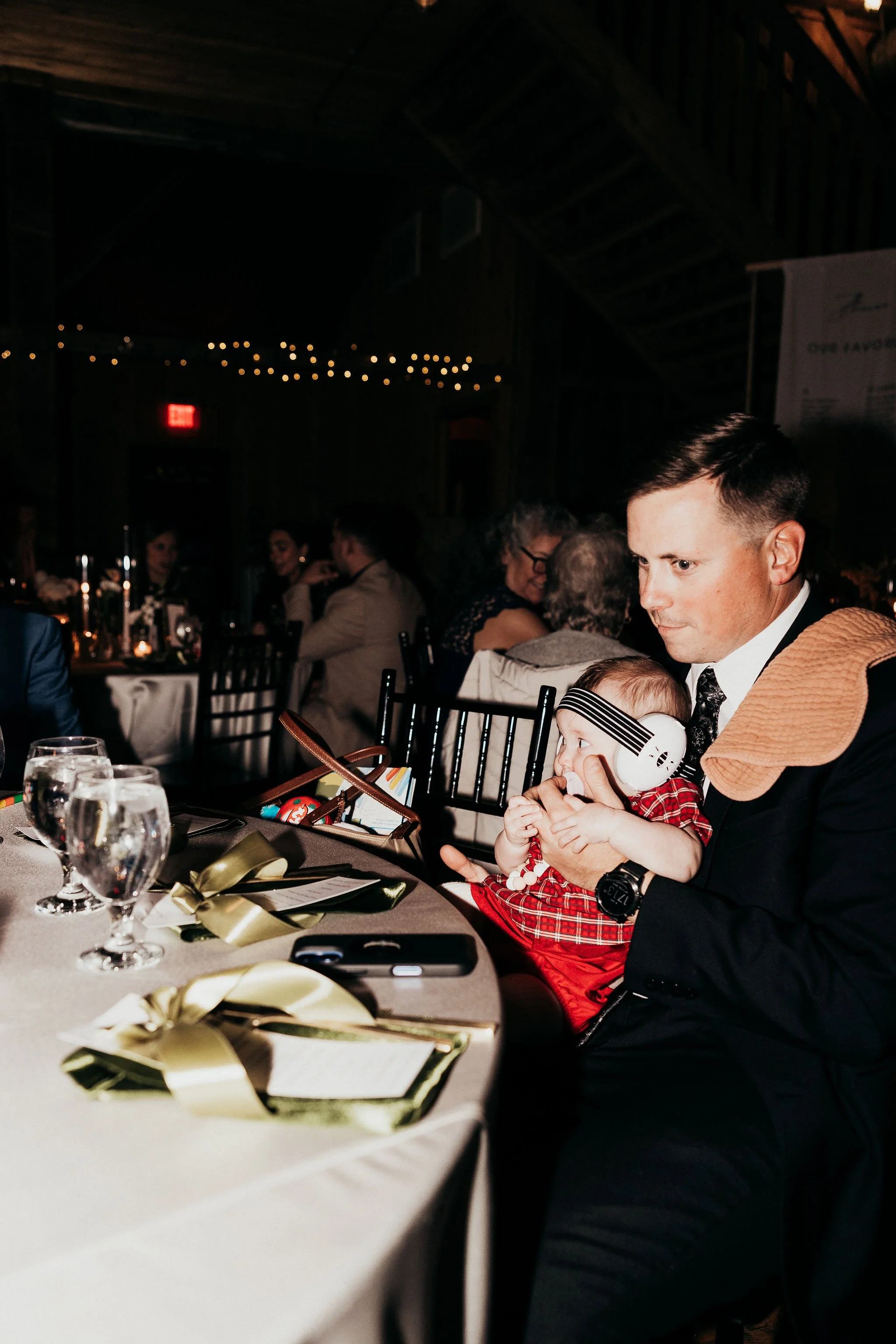 Person holding a young child wearing headphones at a festive indoor event, with decorated tables and string lights in the background.