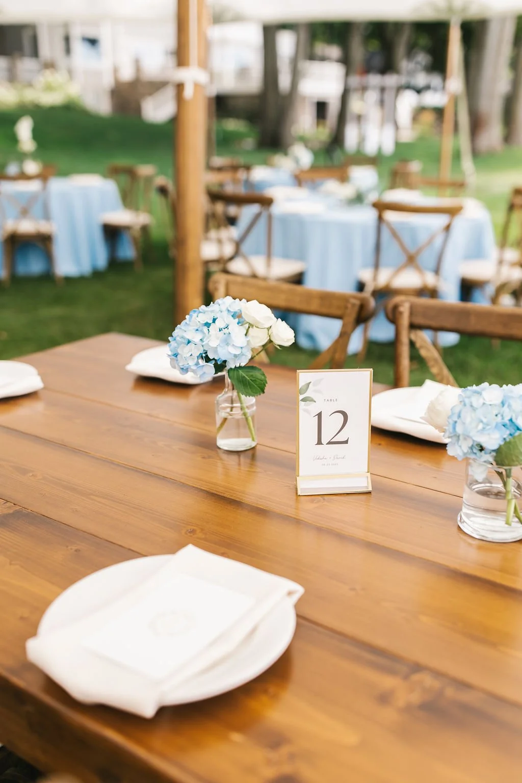 Outdoor wedding reception table decorated with blue and white hydrangea flowers in glass vases, with table number 12 card, white napkins, and wooden chairs, set on green grass with additional tables and chairs in the background.