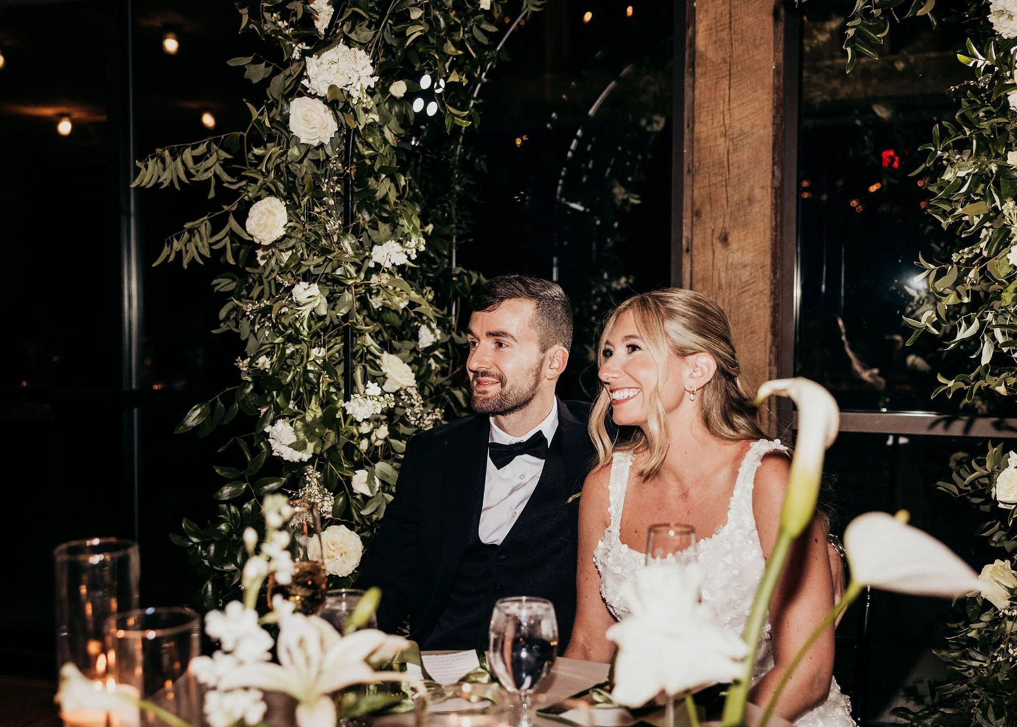 A bride and groom sitting at a decorated wedding table, smiling, with greenery and white flowers surrounding them in a dimly lit indoor setting.