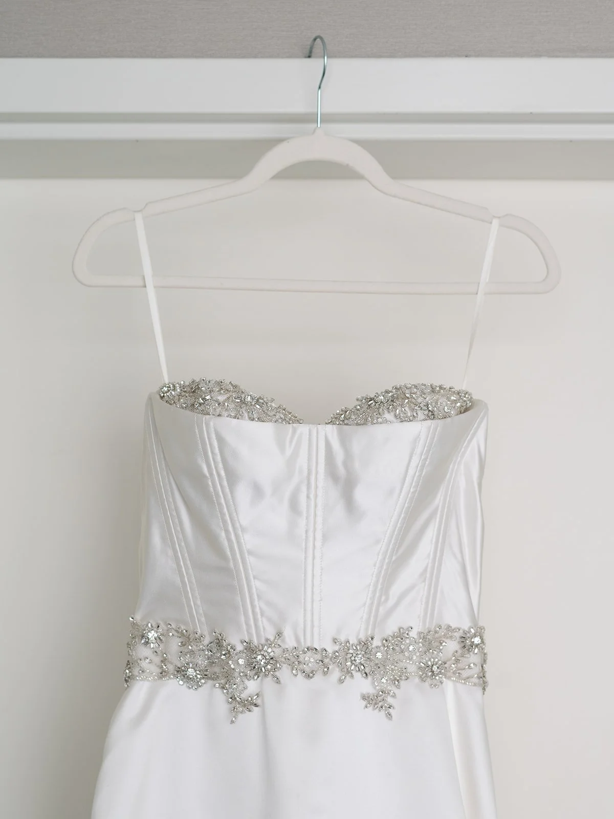 A white wedding dress with beadwork and jeweled details hanging on a white hanger against a plain background.