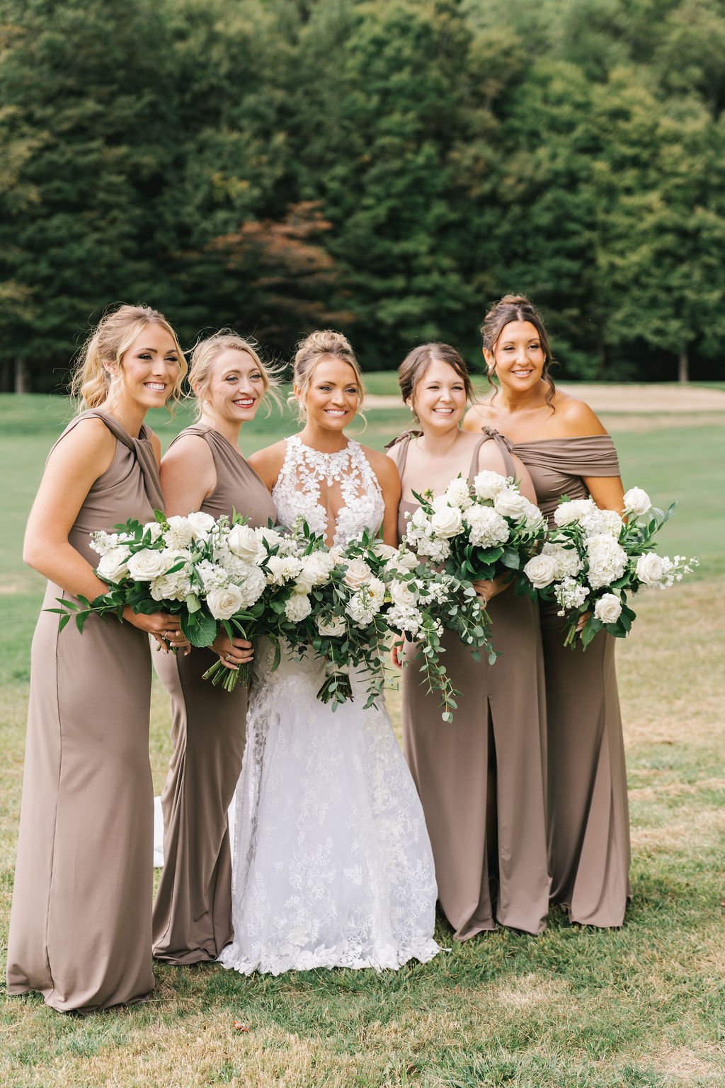 A bride in a white lace wedding dress with five bridesmaids in taupe dresses holding large bouquets of white flowers, standing outdoors on grass with green trees in the background.