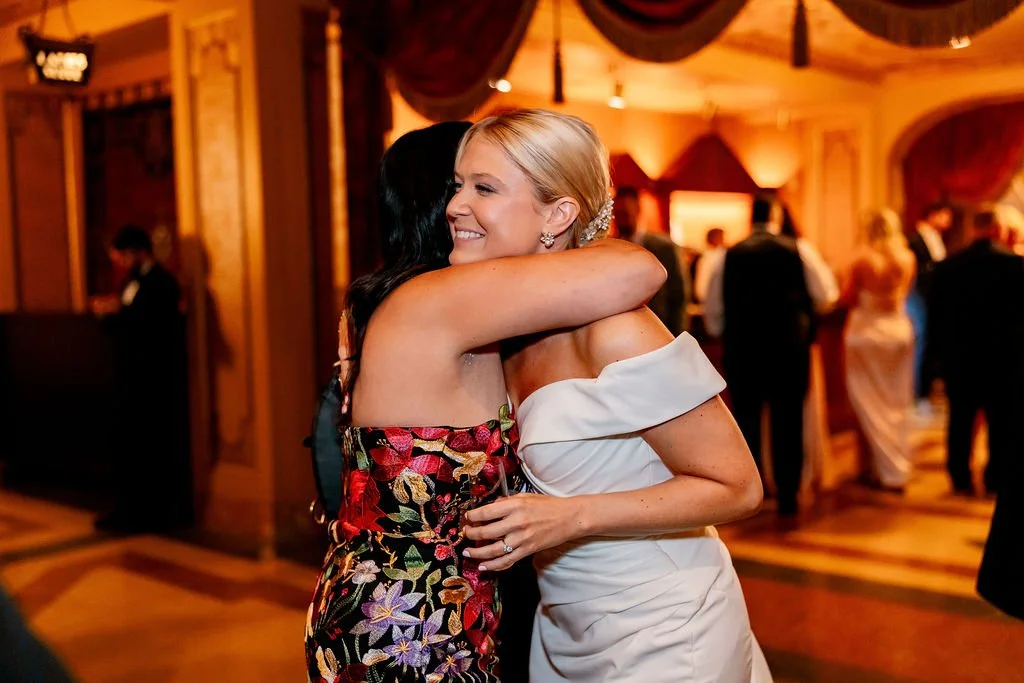 Two women hugging at a formal event, one in a colorful floral dress and the other in an off-the-shoulder white dress, in a warmly lit banquet hall.