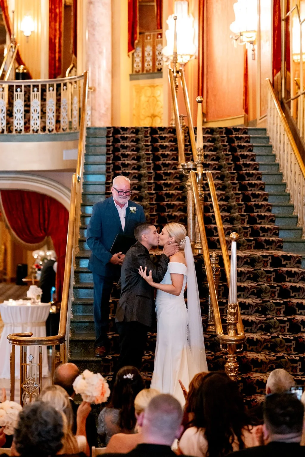 A wedding ceremony taking place on a grand staircase in an elegant, ornate venue. The bride and groom are kissing, and the officiant stands behind them. Guests are seated in front, watching and celebrating.