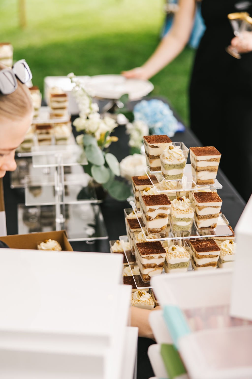 Assorted layered desserts in small glasses arranged on a tiered display at an outdoor event.
