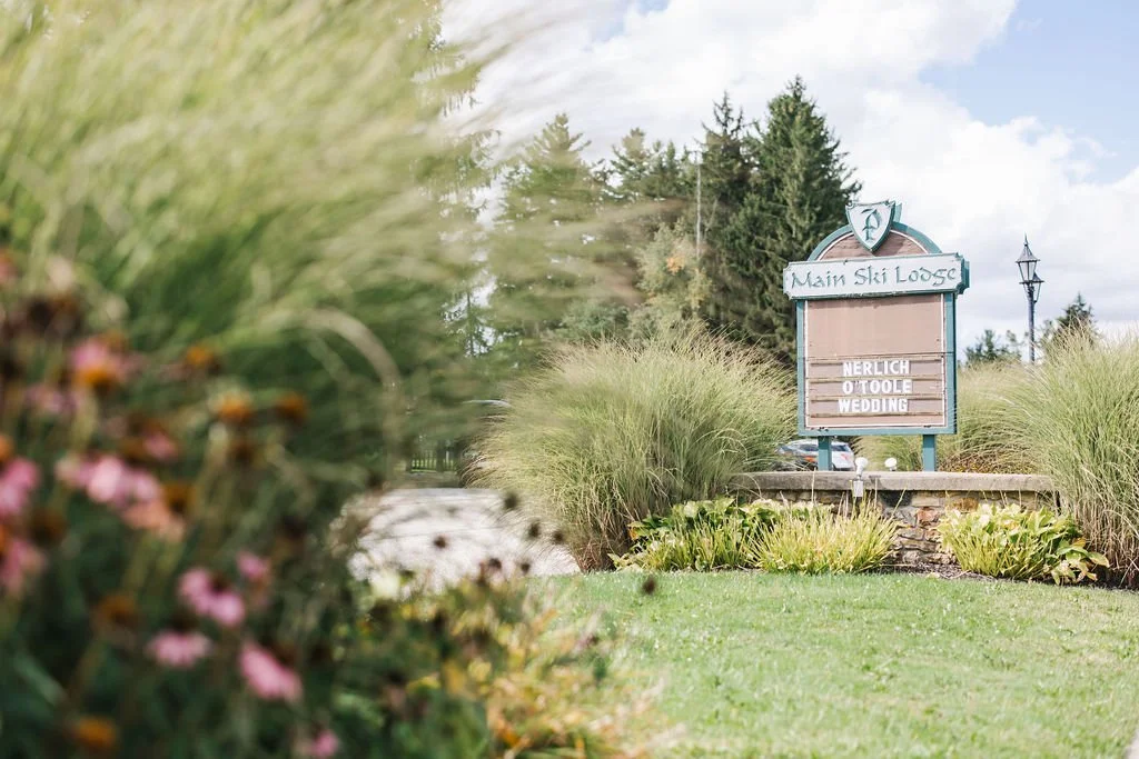 Sign in front of a landscaped area with grass, plants, and ornamental grasses, reading 'Main Ski Lodge' and a schedule for events. Trees and a cloudy sky in the background.