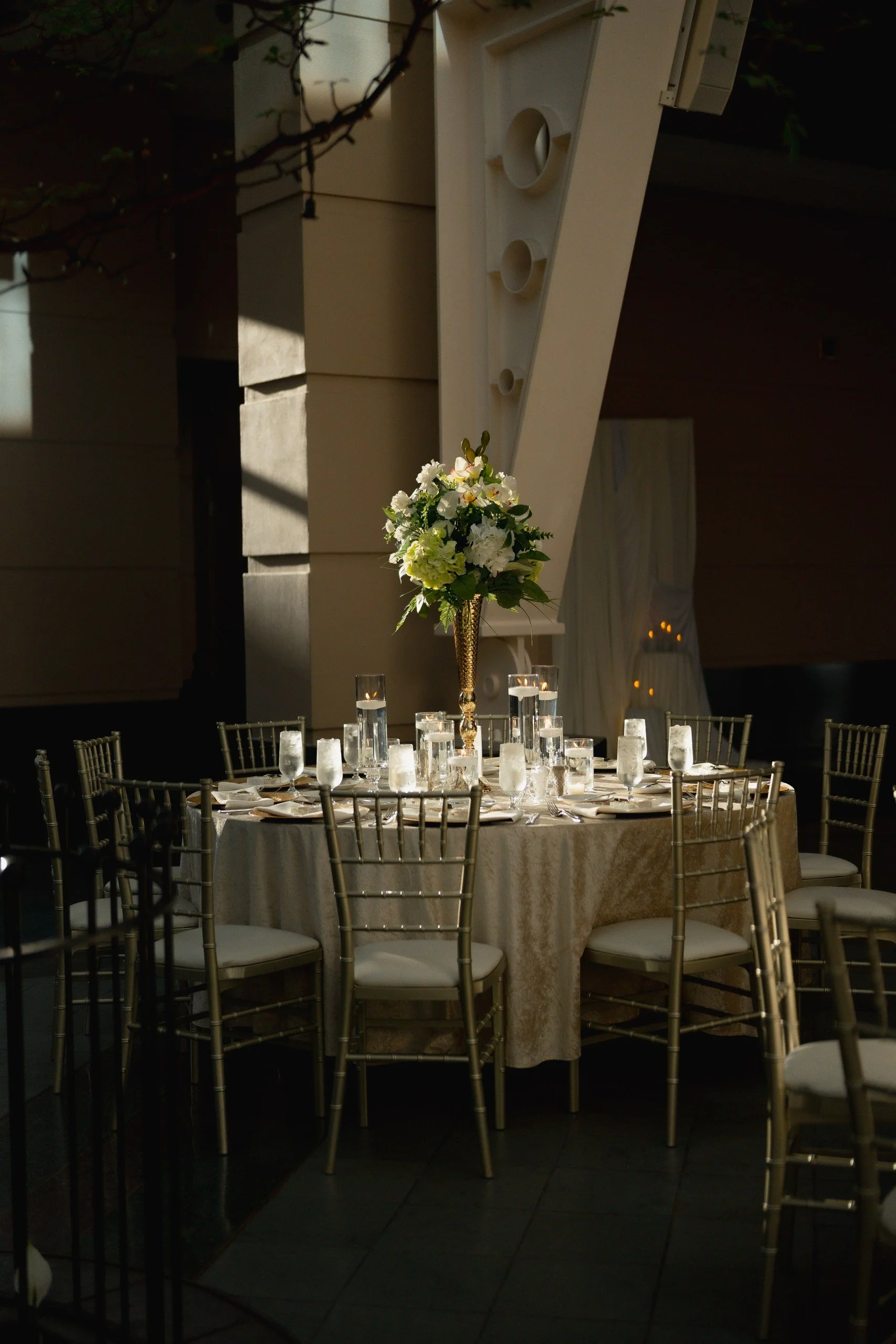 Elegant dining table set for an event with a large floral centerpiece and multiple lit candles, surrounded by gold chairs with white cushions, under warm lighting in a decorated indoor venue.