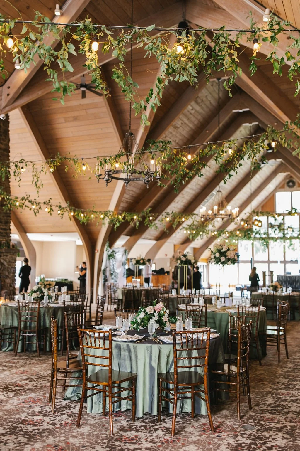 Elegant banquet hall with tables decorated with green tablecloths, floral centerpieces, and set with glassware and silverware. The ceiling features wooden beams, greenery garlands, and hanging string lights, creating a warm, festive atmosphere.
