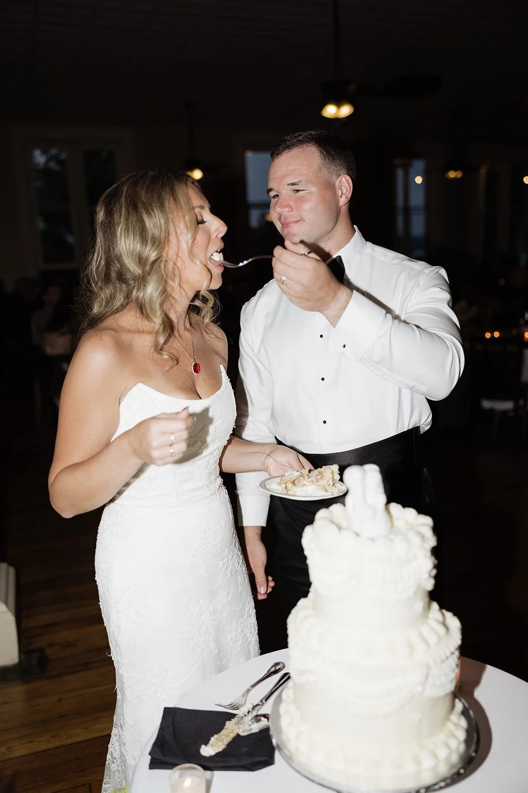 Bride and groom sharing wedding cake at their reception, with a three-tier white wedding cake in the foreground.