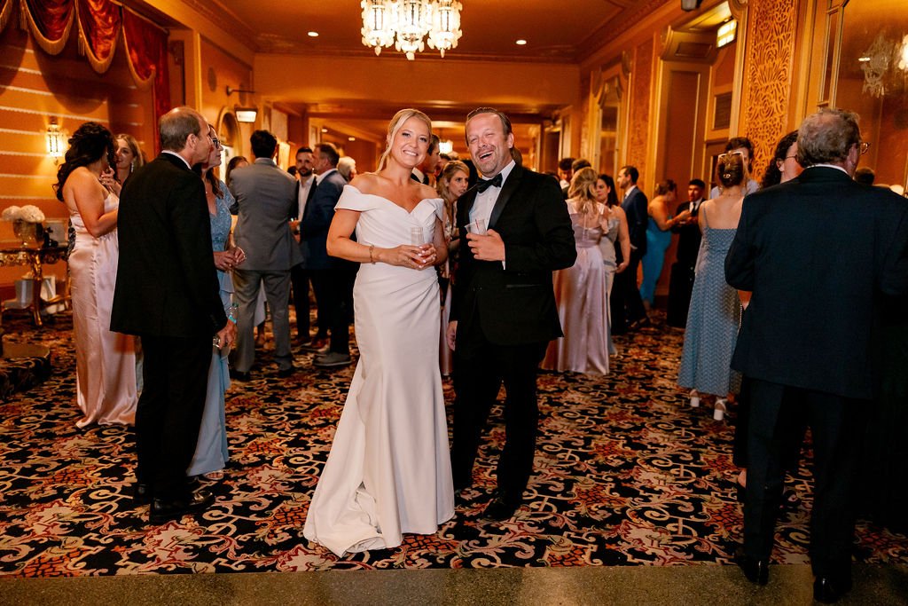 A woman in a white off-shoulder gown and a man in a black tuxedo with a bow tie are smiling and holding drinks at a formal event. Other guests in formal attire are mingling in a ballroom with ornate decor and patterned carpet.