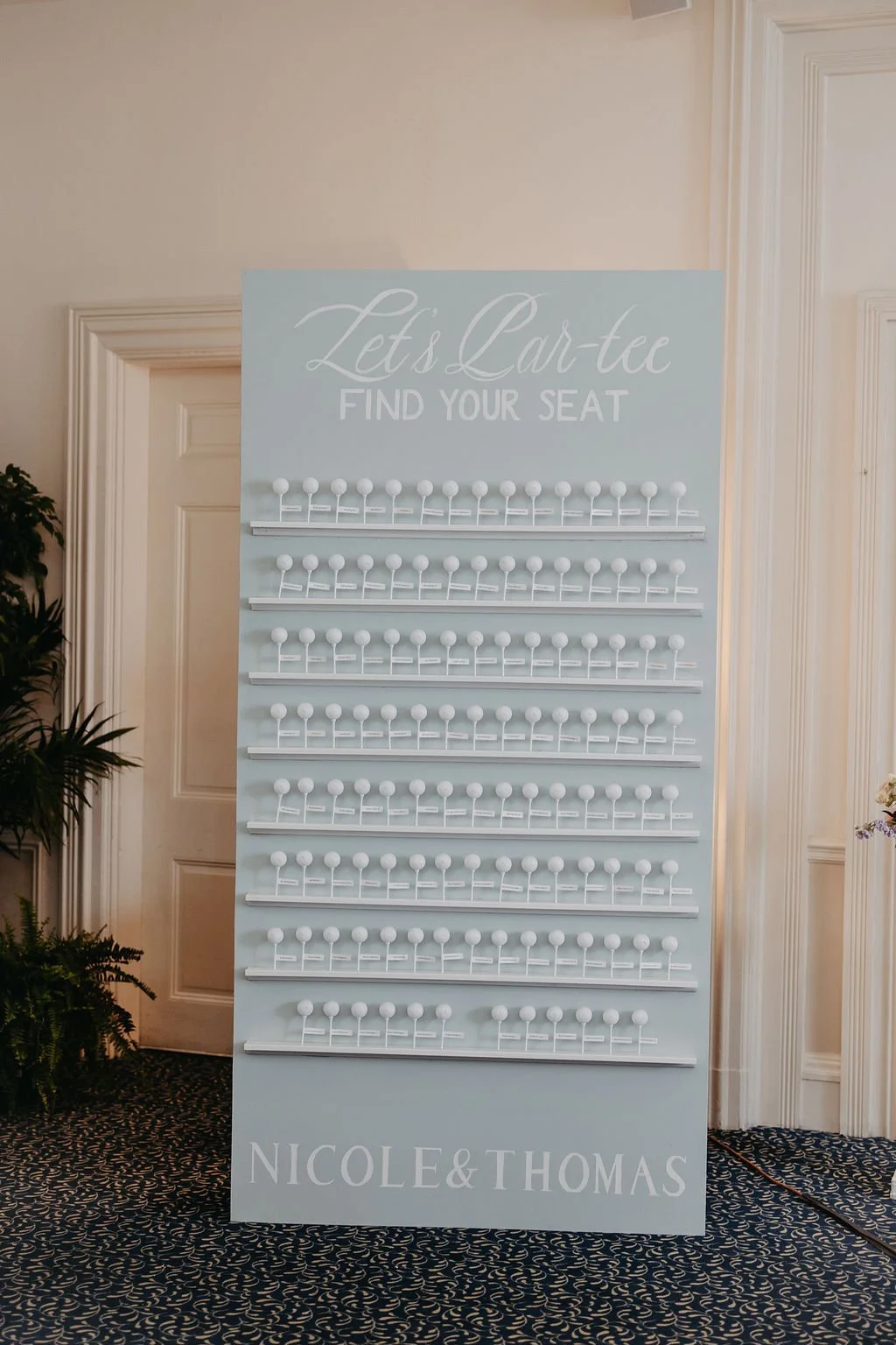 Light blue card display board with the words "Let's Car-tee, Find Your Seat" at the top and rows of small white hooks underneath for holding cards or tags. The bottom of the board has the names "Nicole & Thomas".