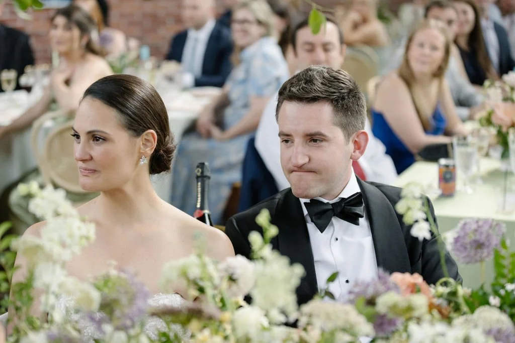 A bride and groom sitting at a wedding reception surrounded by floral decorations, with guests in the background.