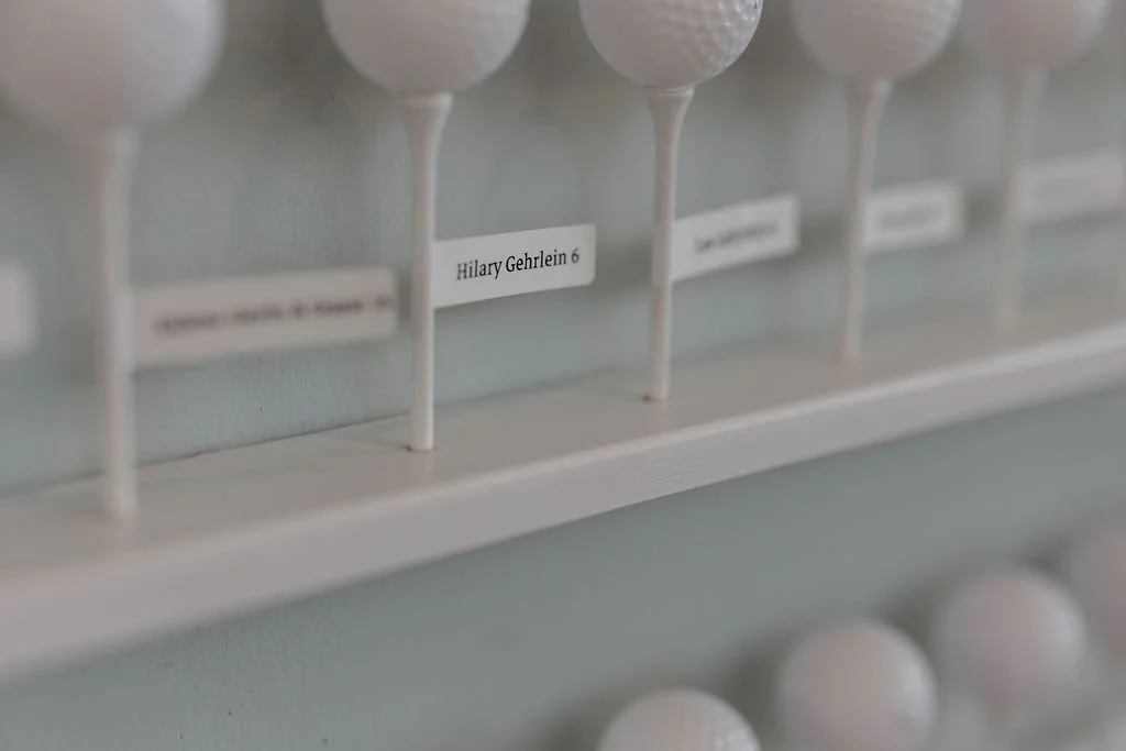 Golf balls arranged in a rack with a label reading "Hilary Gehrlein 6" visible in the background.