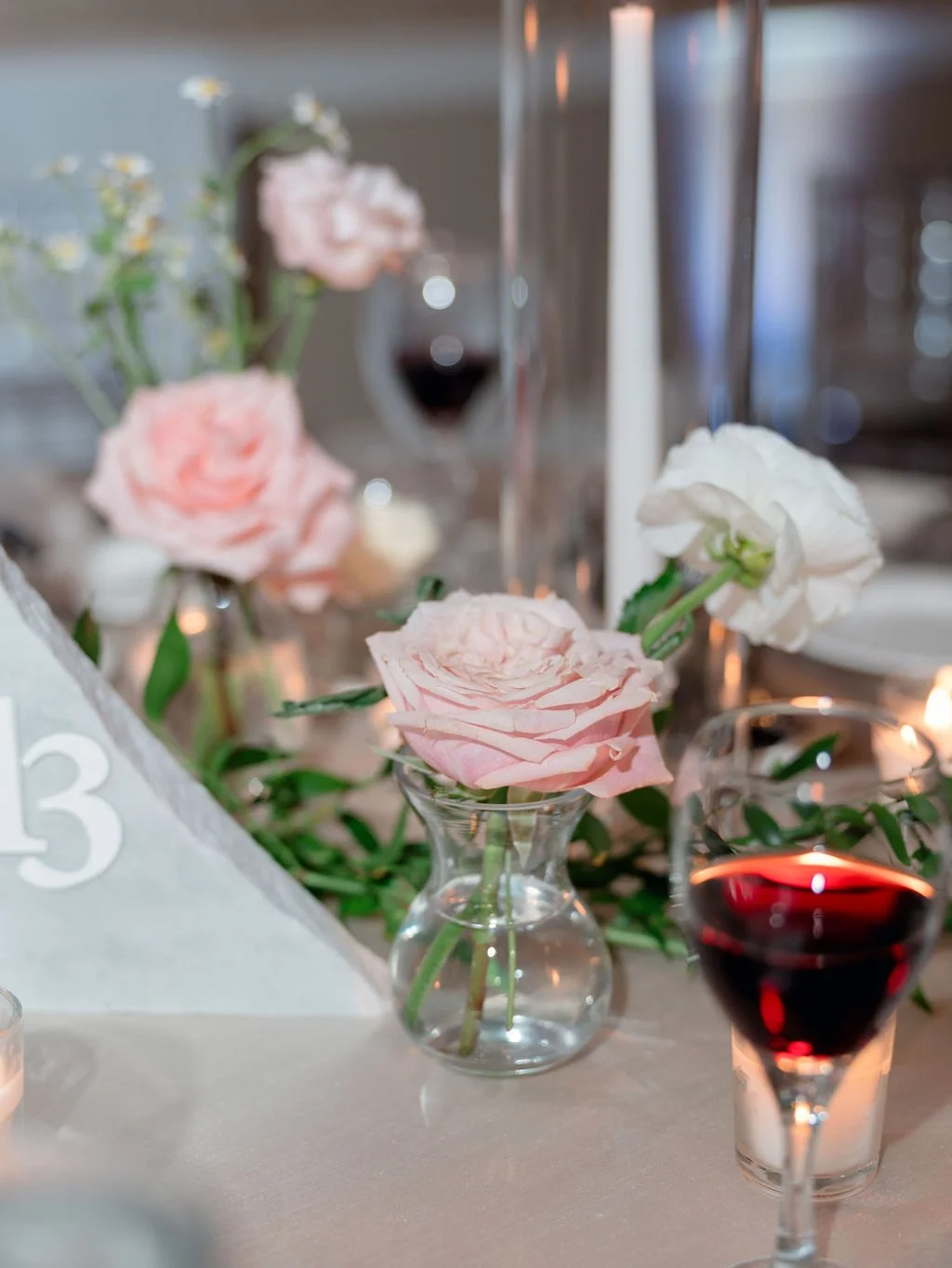 A floral centerpiece with pink and white roses in small glass vases on a table, accompanied by a glass of red wine and candlelight.