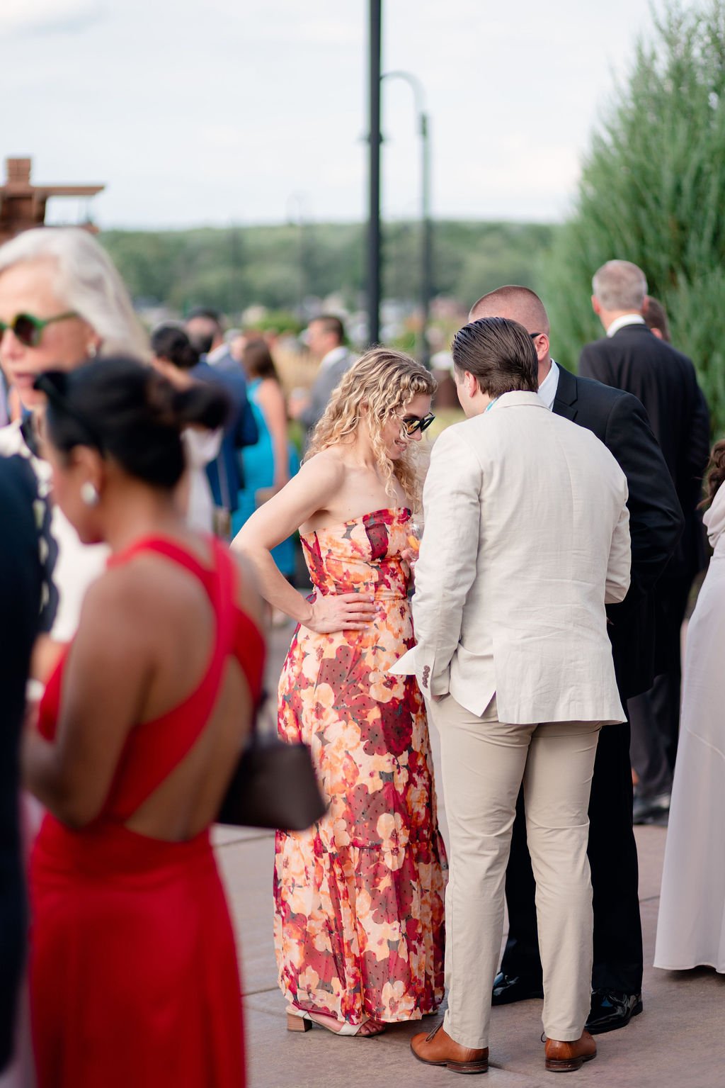 Group of people dressed in formal attire, socializing outdoors at an event, possibly a wedding or a garden party, on a partly cloudy day with greenery in the background.
