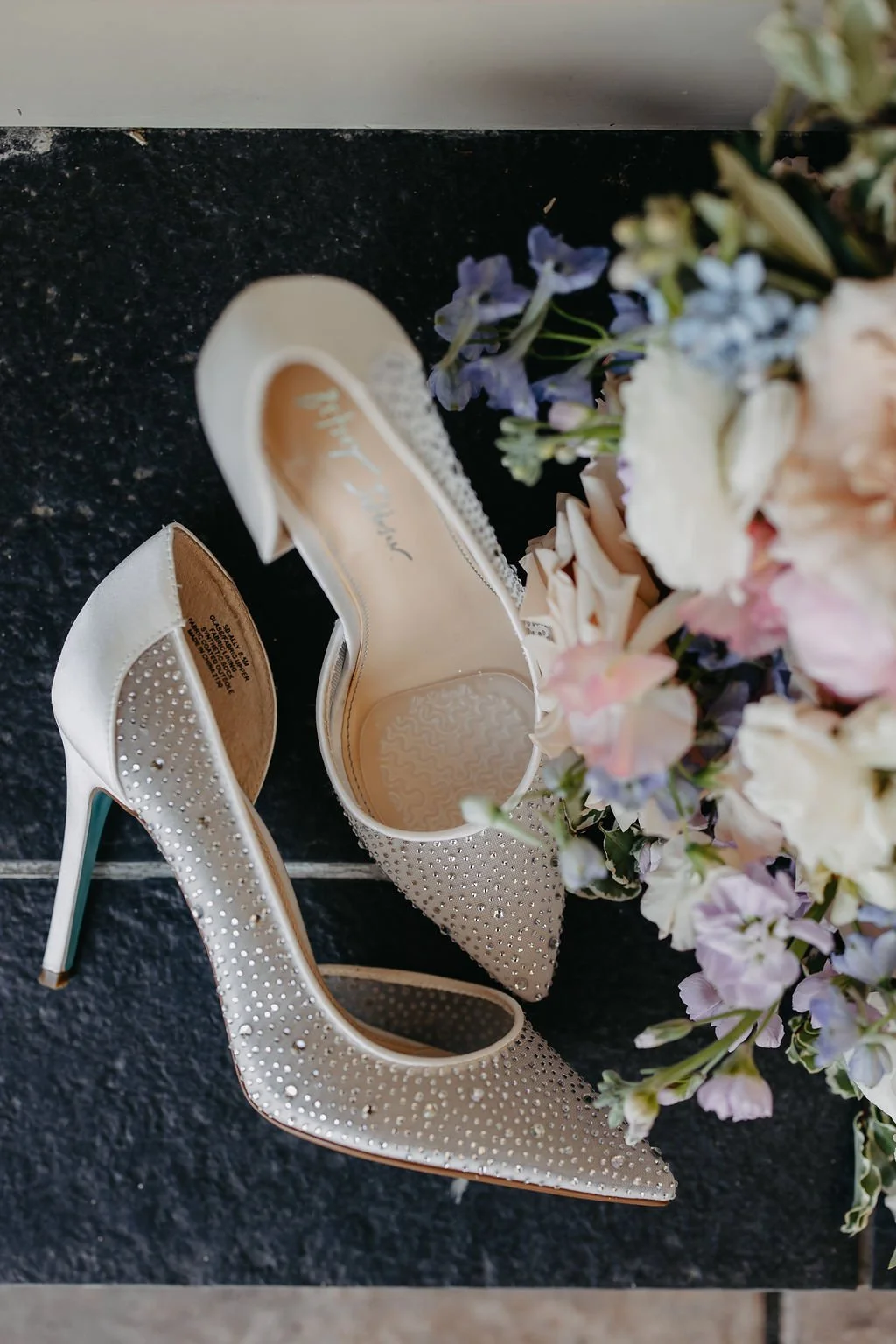 A pair of ivory high-heeled shoes decorated with rhinestones, positioned next to light-colored flowers on a dark surface.