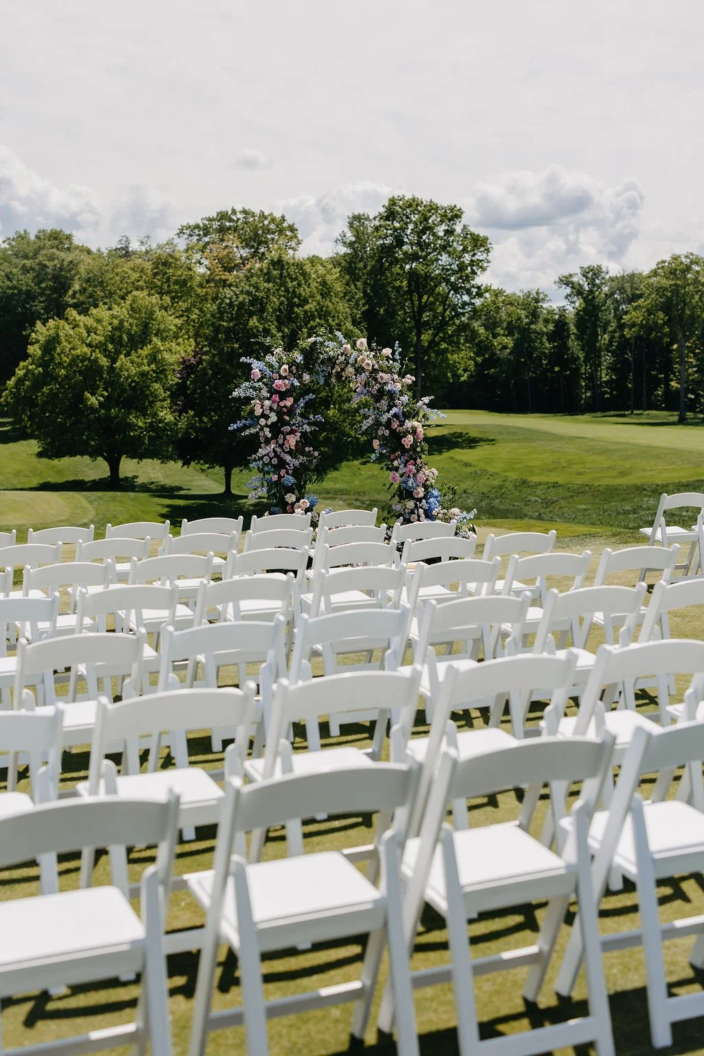 Outdoor wedding setup with white chairs and a floral arch on a grassy field, surrounded by trees and a cloudy sky.