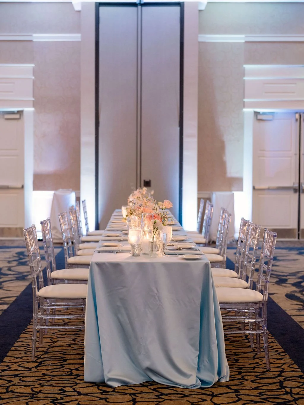 Elegant banquet table set with white tablecloth, pink flowers, candles, and glassware, in a decorated event hall with carpeted floor and wall mirrors.