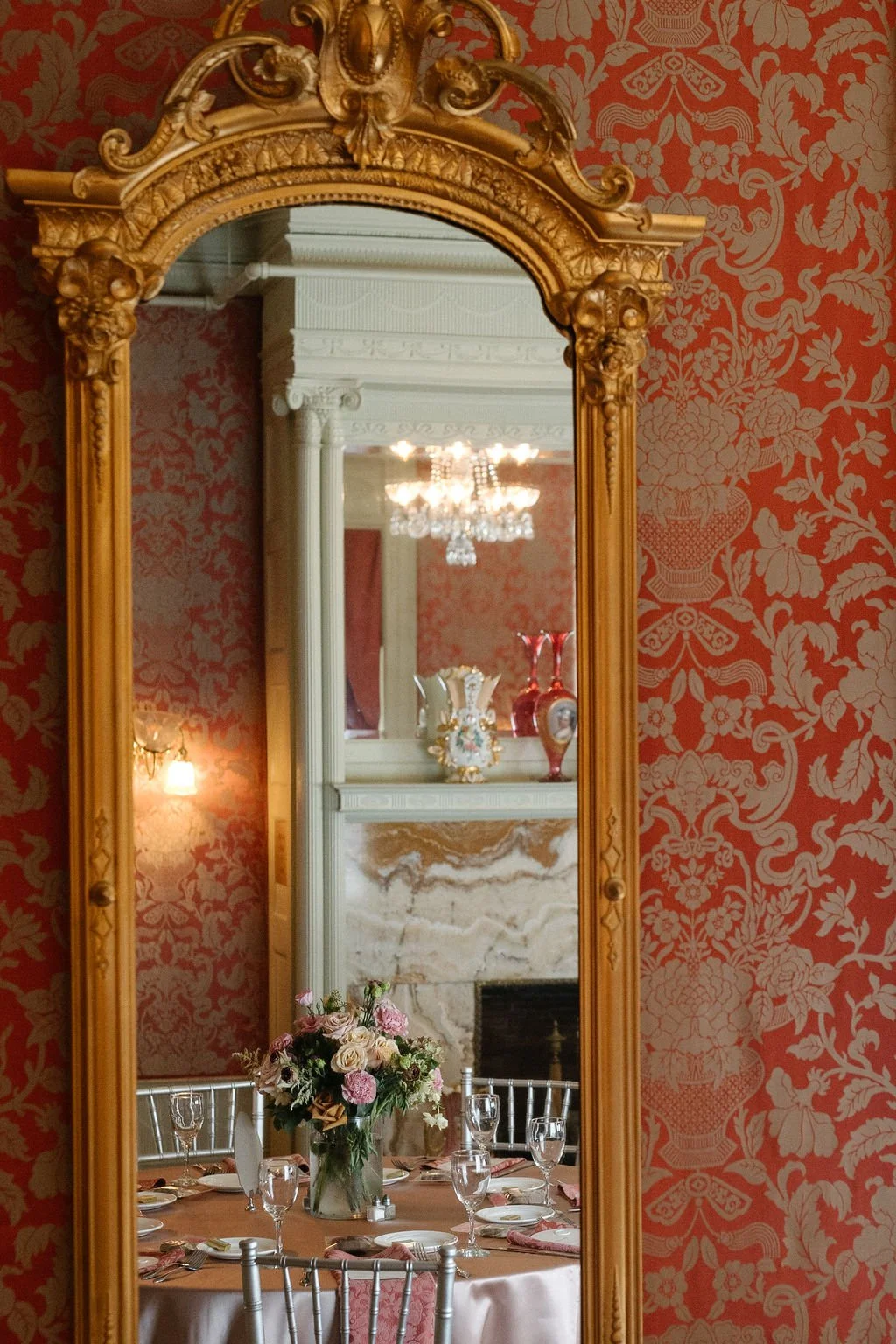 An ornate gold-framed mirror reflecting a decorated dining room with a chandelier, a marble fireplace, a floral centerpiece, and elegant table settings.