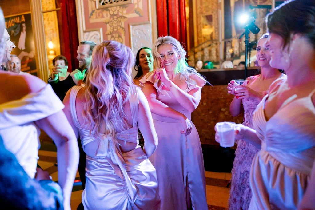Group of women in party dresses dancing and laughing at a social gathering in a decorated venue with warm lighting and wooden accents.